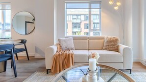 Een moderne woonkamer met een bank, een ronde tafel, een spiegel en een groot raam met uitzicht op gebouwen.