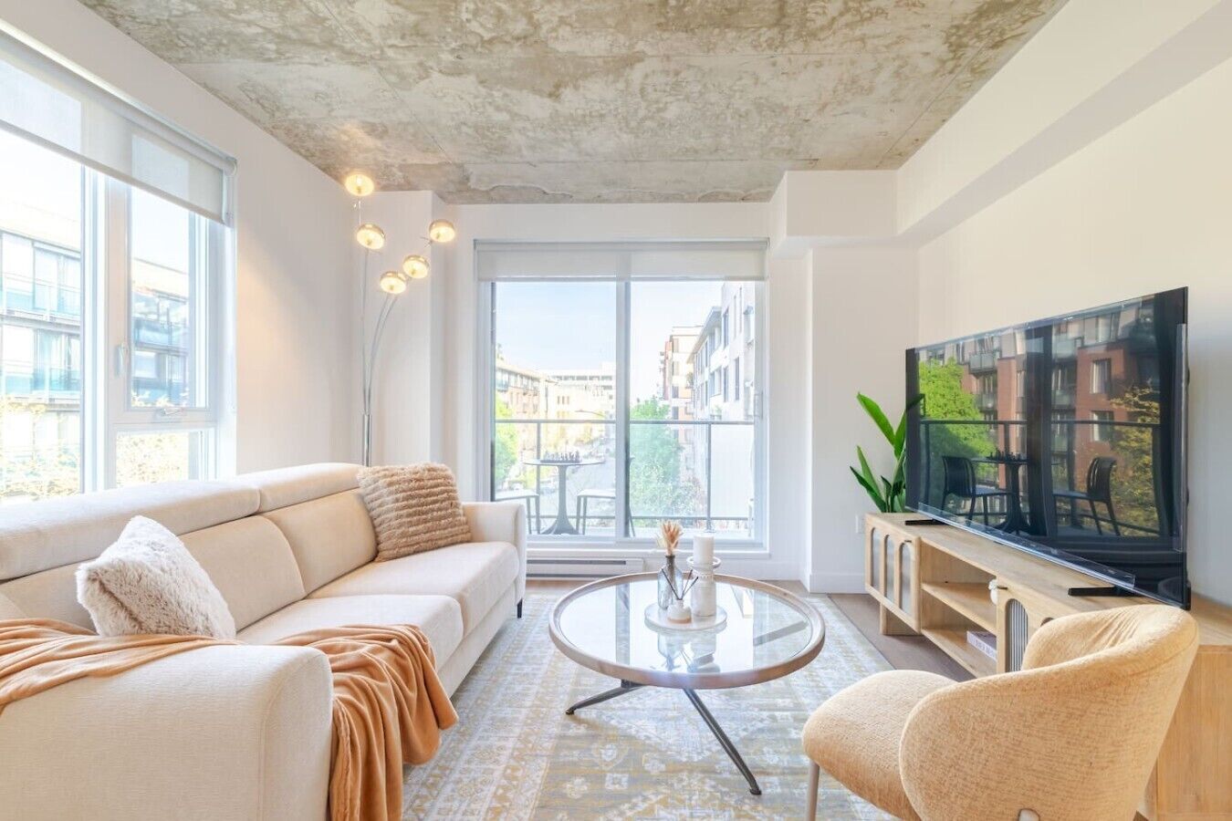 Een moderne woonkamer met een bank, een salontafel, een televisie en een balkon met uitzicht op gebouwen.