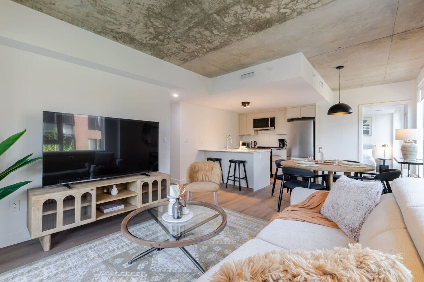 A modern living room with a flat-screen TV, a glass coffee table, a sofa, and a kitchen area with a dining table.