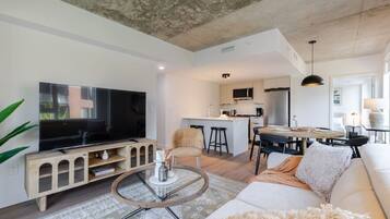 A modern living room with a flat-screen TV, a glass coffee table, a sofa, and a kitchen area with a dining table.