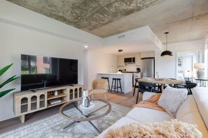 A modern living room with a flat-screen TV, a glass coffee table, a sofa, and a kitchen area with a dining table.