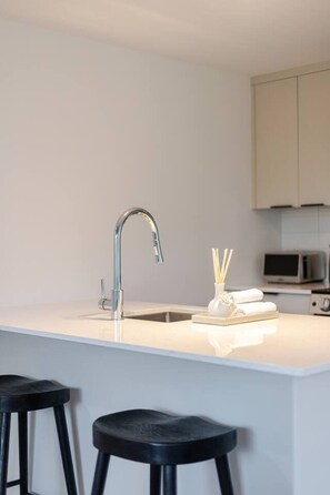 A modern kitchen with a white countertop, a stainless steel faucet, and two black stools. - Grand Horizon Montréal | Free Parking (Montreal)
