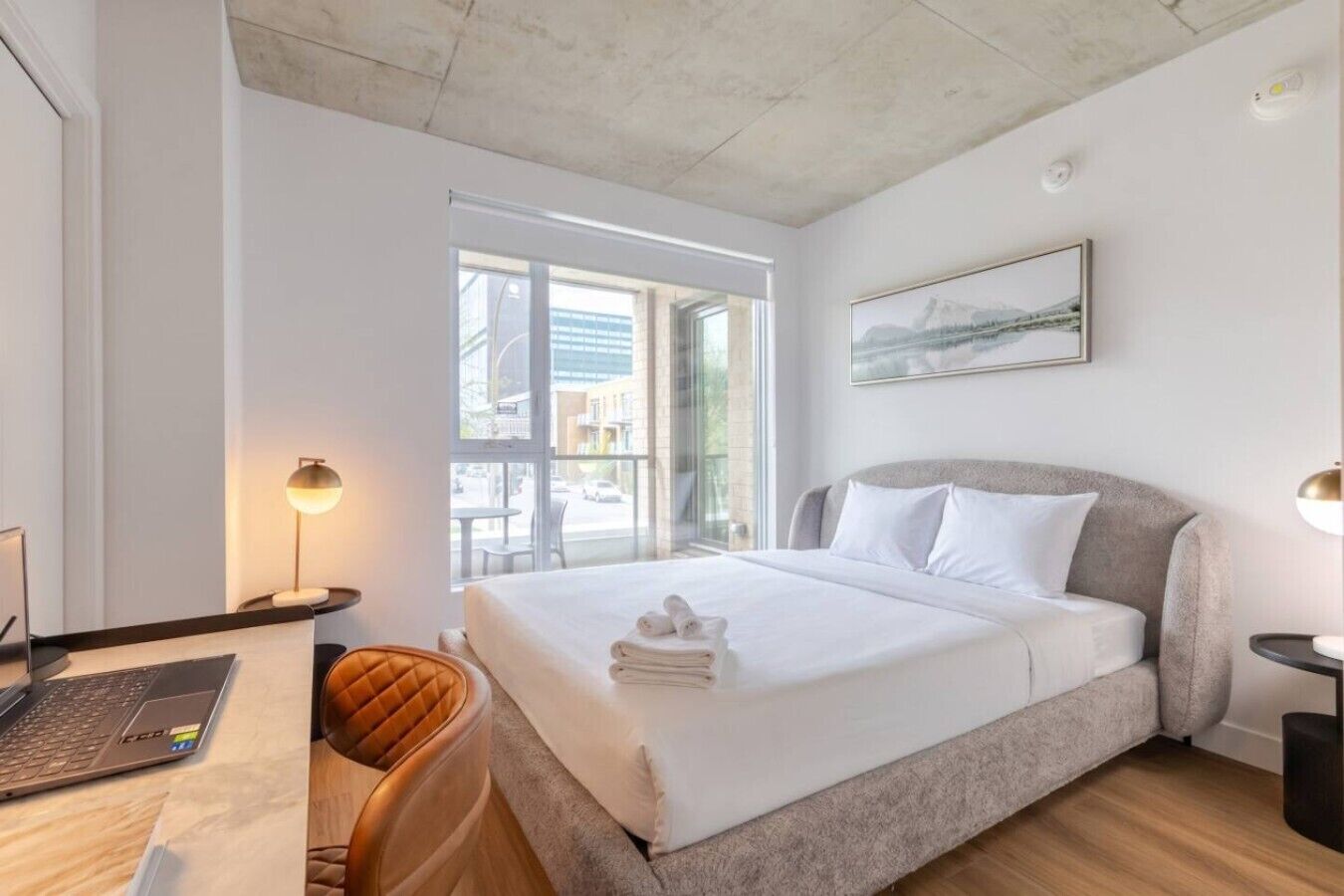 A modern hotel room with a bed, a desk with a laptop, a chair, a window with a view, and a painting on the wall.