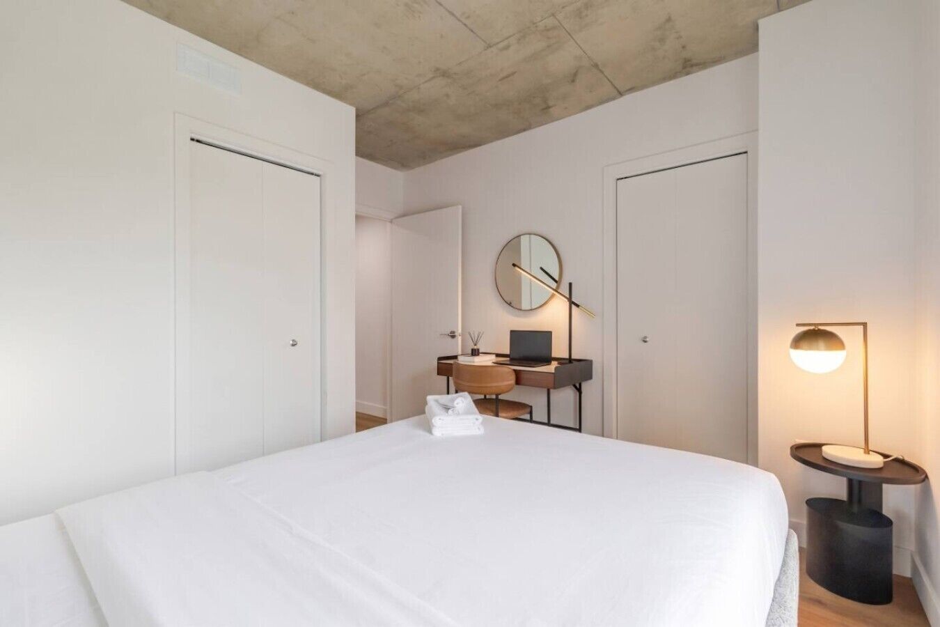 A bedroom with a bed, a desk with a laptop, a round mirror, and a bedside lamp.