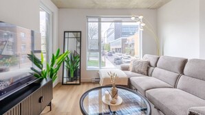Ein modernes Wohnzimmer mit einer Couch, einem Glastisch, einem Fernseher, einem großen Fenster und einer Pflanze.