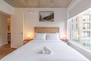 A hotel room with a large bed, two bedside tables with lamps, a painting on the wall, and a view through a window. - CityGlow Montréal w/ Free Parking (Montreal)