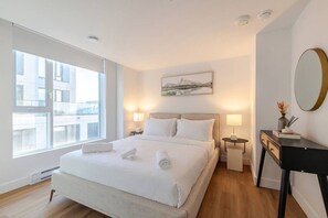 A modern hotel room with a large bed, bedside lamps, a small table with a mirror, and a window with a city view.