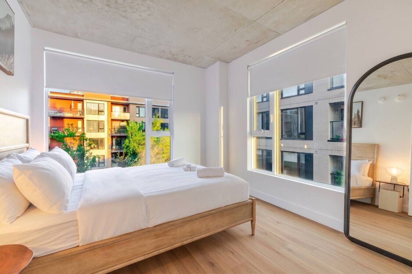 Een moderne slaapkamer met een houten bed, grote ramen en uitzicht op een gebouw met balkons.
