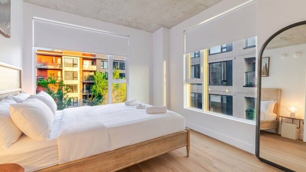 A modern bedroom with a wooden bed, large windows, and a view of a building with balconies. - CityGlow Montréal w/ Free Parking (Montreal)
