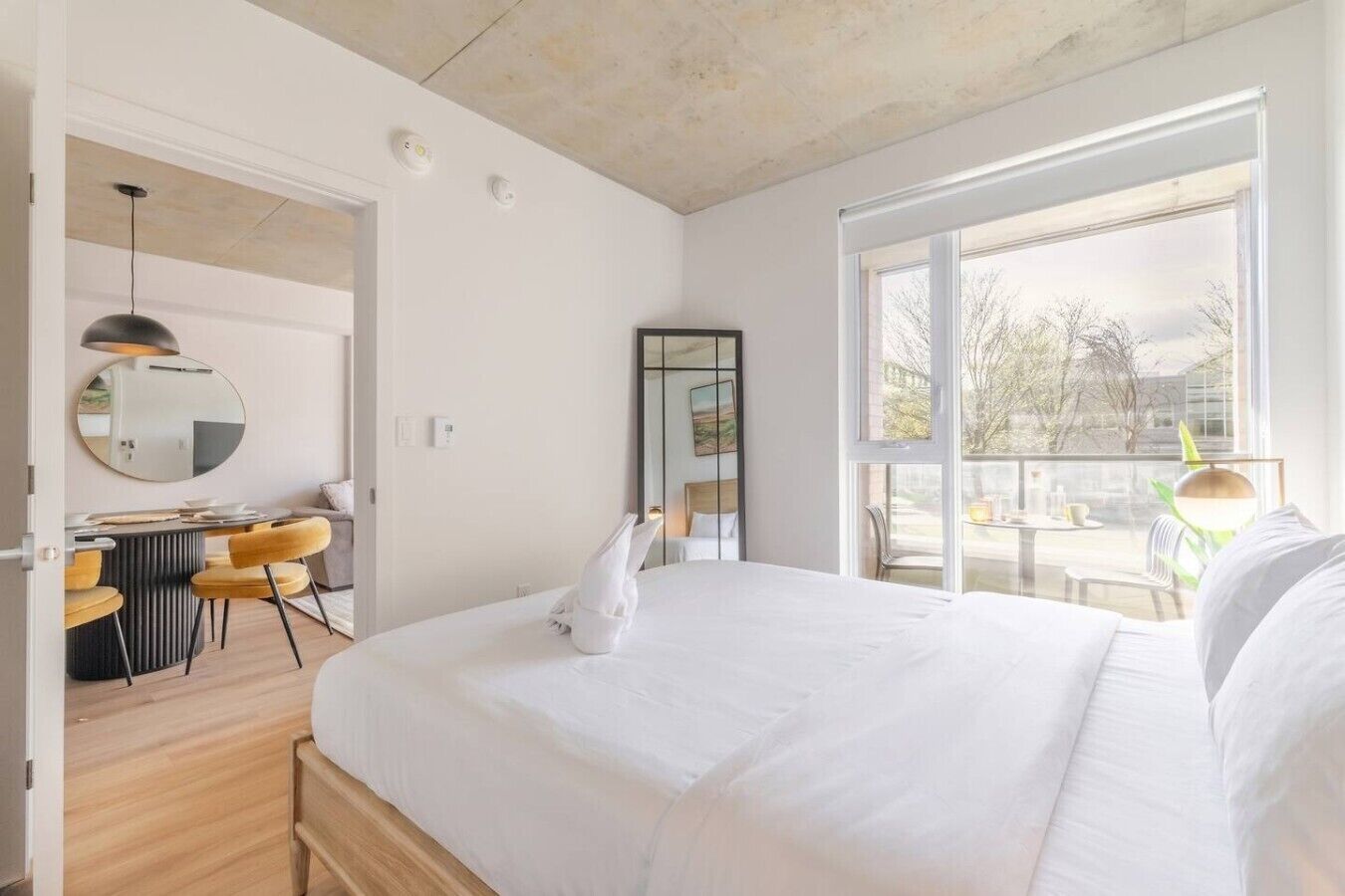 A modern hotel room with a large bed, a dining area with a round table and chairs, a large window with a view, and a balcony.