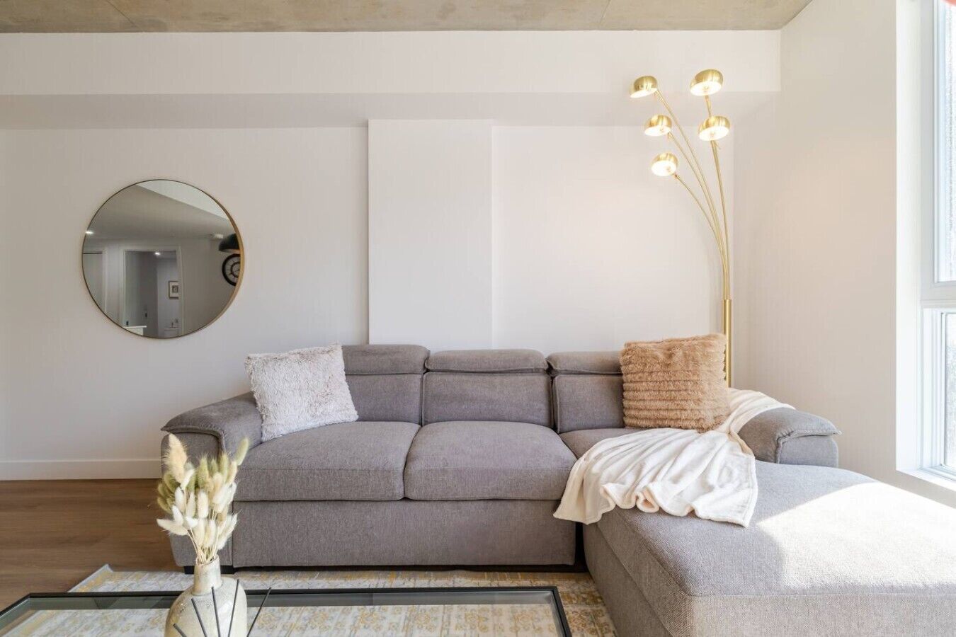 Een moderne zithoek met een grijze modulaire bank, een ronde spiegel, een vloerlamp en een vaas met gedroogde bloemen.