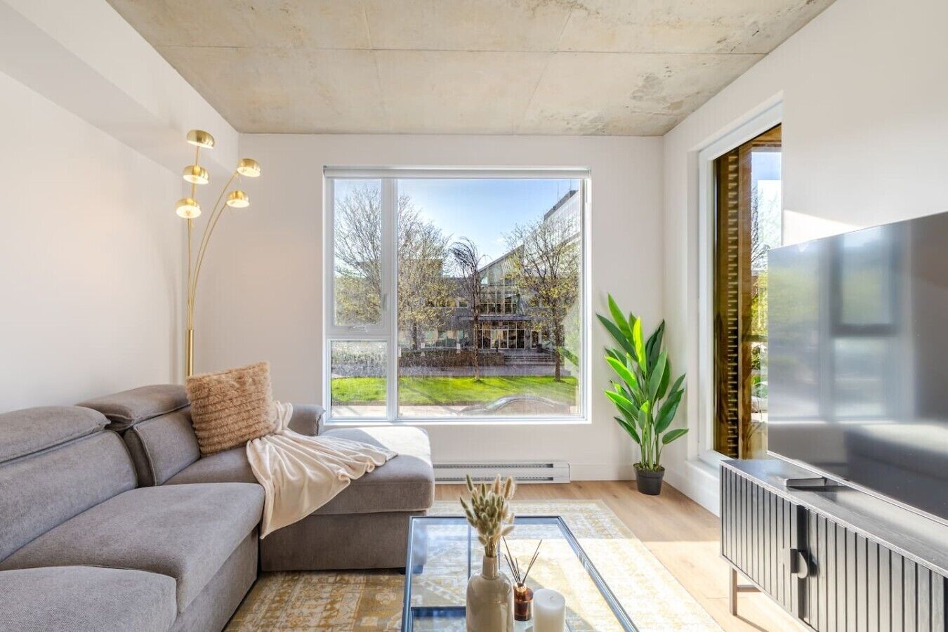Een moderne woonkamer met een grijze bank, een glazen salontafel, een groot raam met uitzicht op een huis en bomen, en een flatscreen-televisie.
