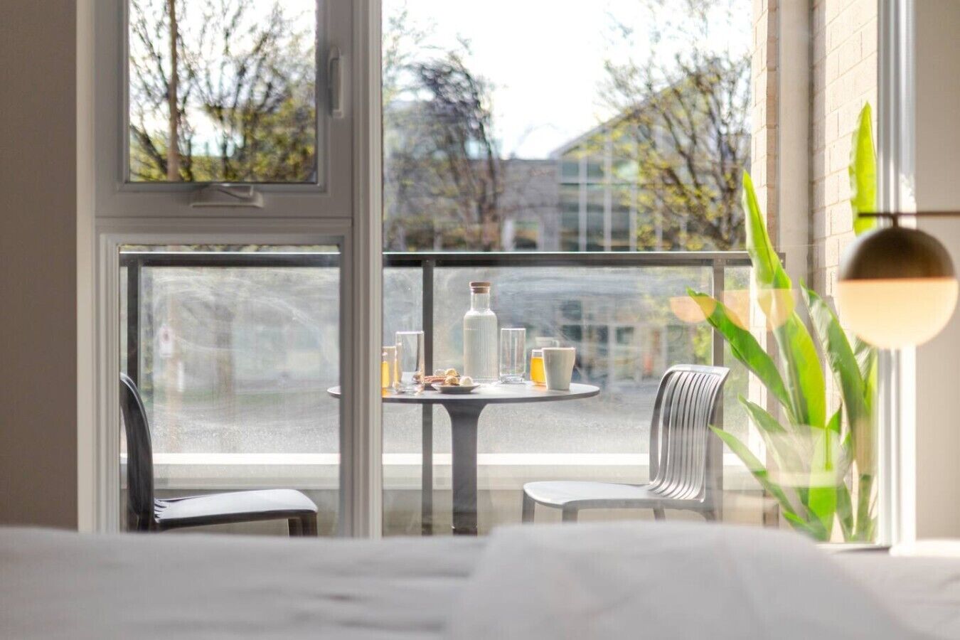 A room with a bed, a balcony with a table and chairs, and a view of trees and buildings.