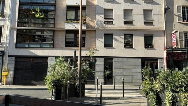 Un immeuble à plusieurs étages doté d’une architecture moderne, avec un mélange de balcons et de grandes fenêtres, et une entrée au niveau du sol avec une devanture visible.