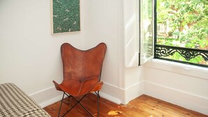 A room with a wooden floor, a brown leather chair, a bed with a checkered bedspread, a window with a view of greenery, and a wall with a green artwork. - Cheese&Wine™ | Amoreiras Garden 3 (Lisbon)