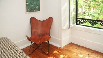 Une pièce avec un plancher en bois, un fauteuil en cuir marron, un lit recouvert d’une couverture à carreaux, une fenêtre donnant sur un paysage verdoyant et un mur orné d’une œuvre d’art verte.