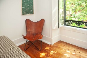 Ein Zimmer mit Holzboden, einem braunen Ledersessel, einem Bett mit kariertem Überwurf, einem Fenster mit Blick auf Grünflächen und einer Wand mit einem grünen Kunstwerk.
