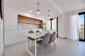 A modern kitchen with white cabinetry, a dining table with chairs, pendant lights, and a view of buildings through a large window. - LXR Indico 2-Bedroom Apartment by the Sea (Fuengirola)