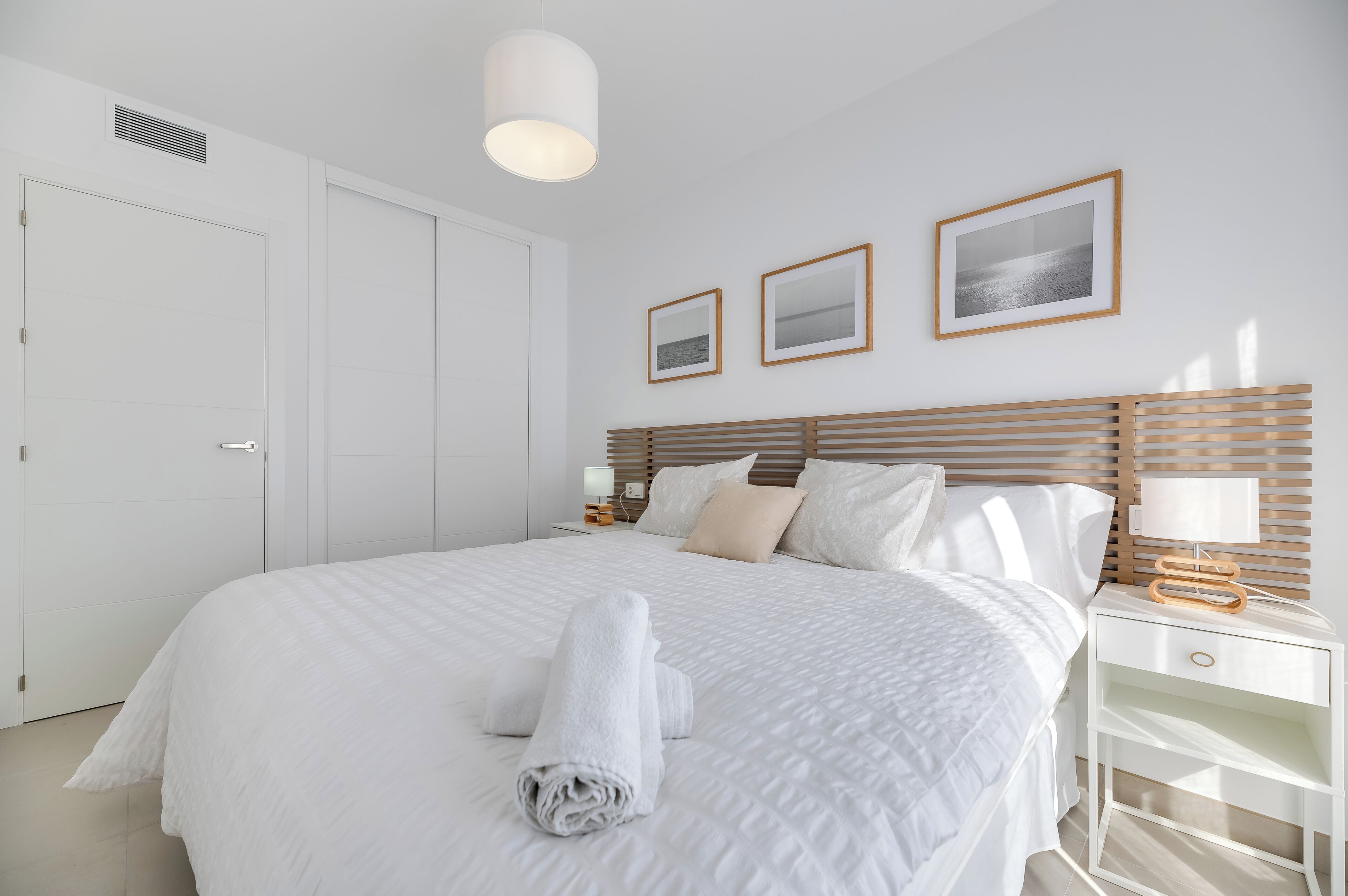 A neatly made bed with white linens and a rolled towel, a bedside table with a lamp, and a wall-mounted picture frame.