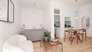 Ein modernes, minimalistisches Schlafzimmer mit Bett, Nachttischen und einem Beistelltisch. Es gibt eine Essecke mit Tisch und Stühlen, und durch eine offene Tür ist eine Küche zu sehen.