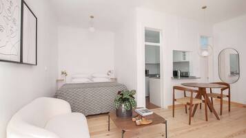 Een moderne, minimalistische slaapkamer met een bed, nachtkastjes en een nachtkastje. Er is een eethoek met een tafel en stoelen, en een keuken is zichtbaar door een open deur.
