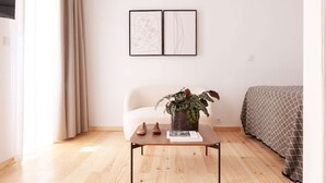 Ein Zimmer mit einem Bett, einem Sessel, einem Tisch mit einer Pflanze und zwei gerahmten Kunstwerken an der Wand.