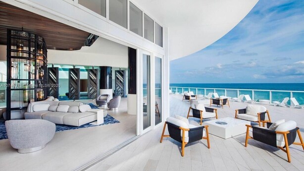 A modern outdoor lounge area with a white sofa, armchairs, and a dining table, overlooking the ocean.