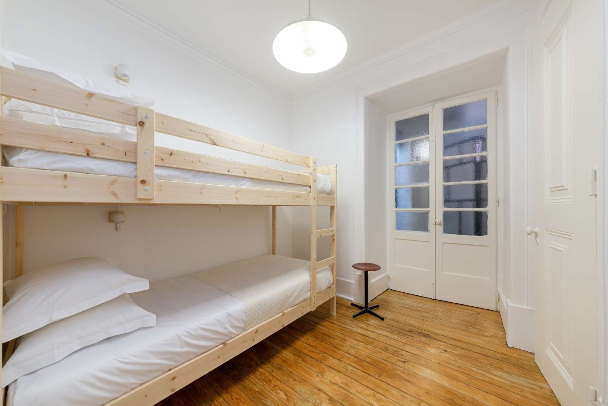 Une chambre avec des lits superposés, un plancher en bois, des murs blancs, un petit tabouret et une porte dont les panneaux sont en verre dépoli.