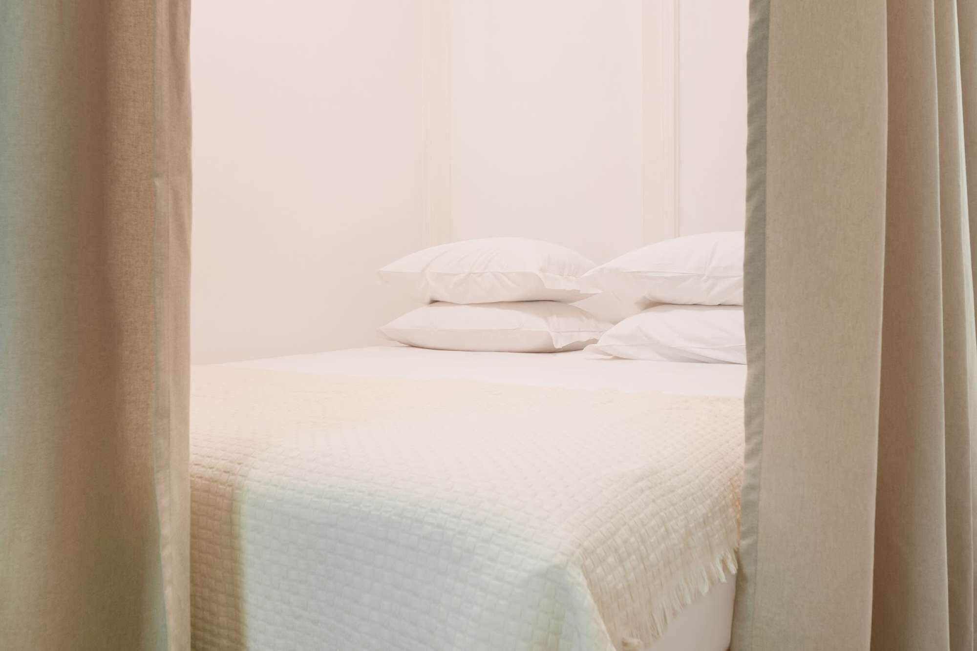 Un llit amb roba de llit i almoinos blancs, cortines entreobertes, i una paret sense decoració al fons.