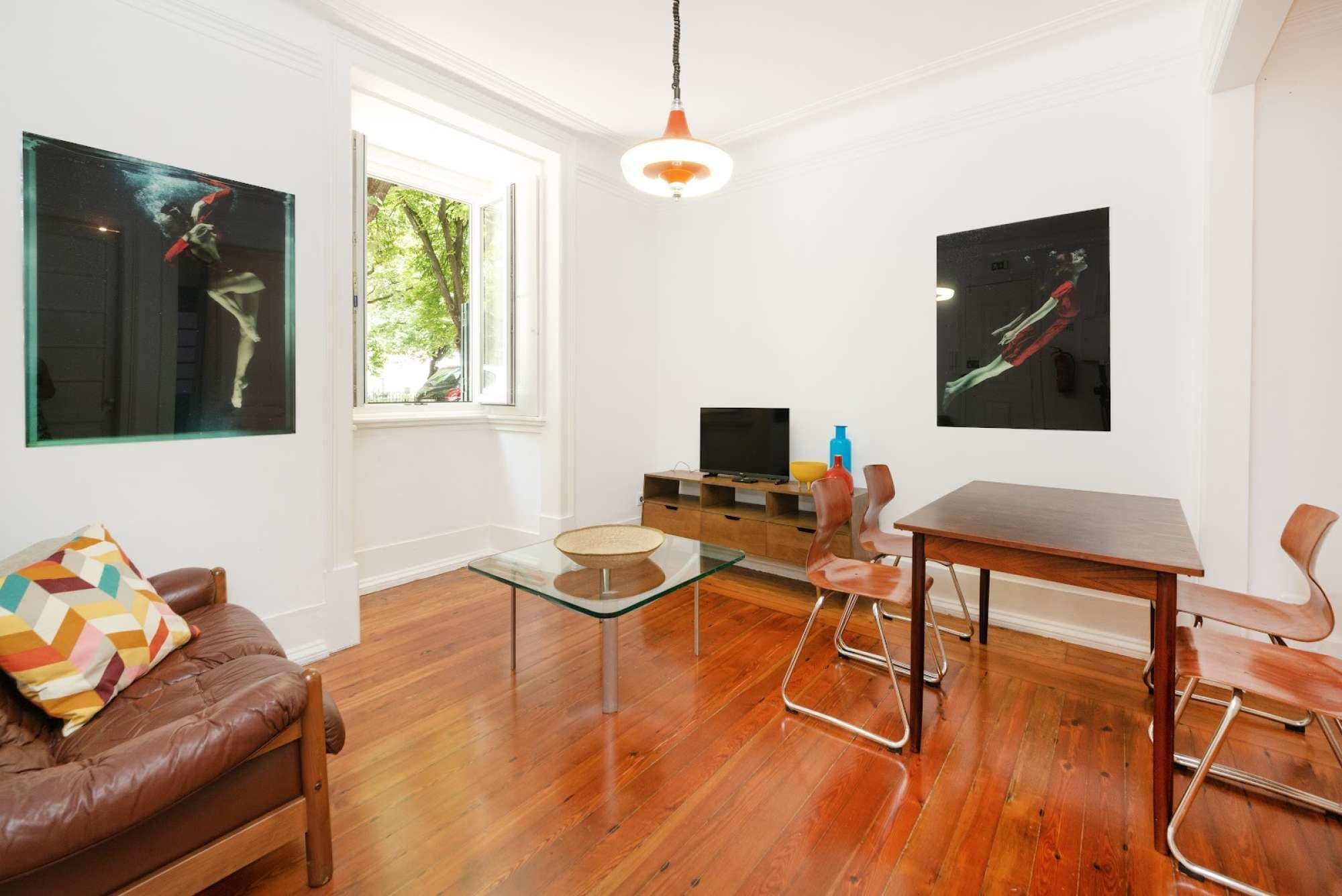 Un salon moderne avec un plancher en bois, une table basse en verre, un canapé en cuir marron, une table à manger avec des chaises en métal et des œuvres d’art accrochées aux murs.