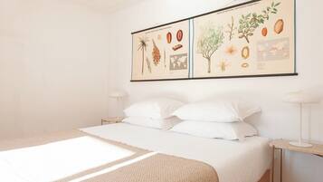 Un dormitorio con cama, ropa de cama blanca, una ilustración botánica enmarcadas en la pared y lámparas de noche.