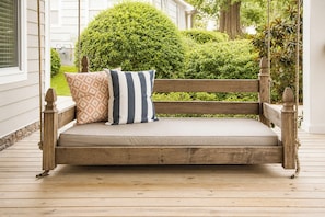 A wooden porch swing with cushions, set on a wooden deck with a view of a well-maintained hedge and a house in the background. - The Retreat, 3BR Wlk-SQ+grill by Velvet Ditch Villas (Oxford)