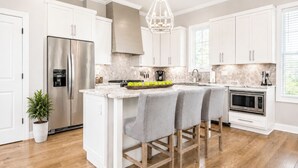 A modern kitchen with white cabinetry, a stainless steel refrigerator, a built-in oven, and a central island with bar stools. - The Retreat, 3BR Wlk-SQ+grill by Velvet Ditch Villas (Oxford)