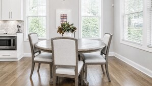 A dining room with a round table, four chairs, a chandelier, and a kitchen area with white cabinets and a microwave. - The Retreat, 3BR Wlk-SQ+grill by Velvet Ditch Villas (Oxford)