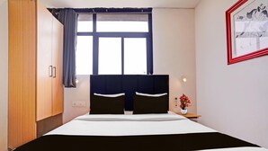 Hotellihuone, jossa on sänky, yöpöytä, verhoilla varustettu ikkuna, seinään kiinnitetty valaisin ja kehystetty kuva.