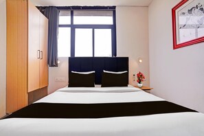 Une chambre d’hôtel avec un lit, une table de chevet, une fenêtre avec des rideaux, un lampadaire fixé au mur et un tableau encadré.