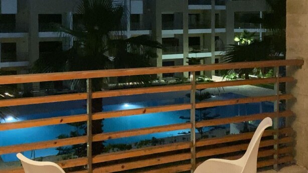 A balcony with two white chairs, a wooden floor, and a view of a swimming pool and buildings at night.