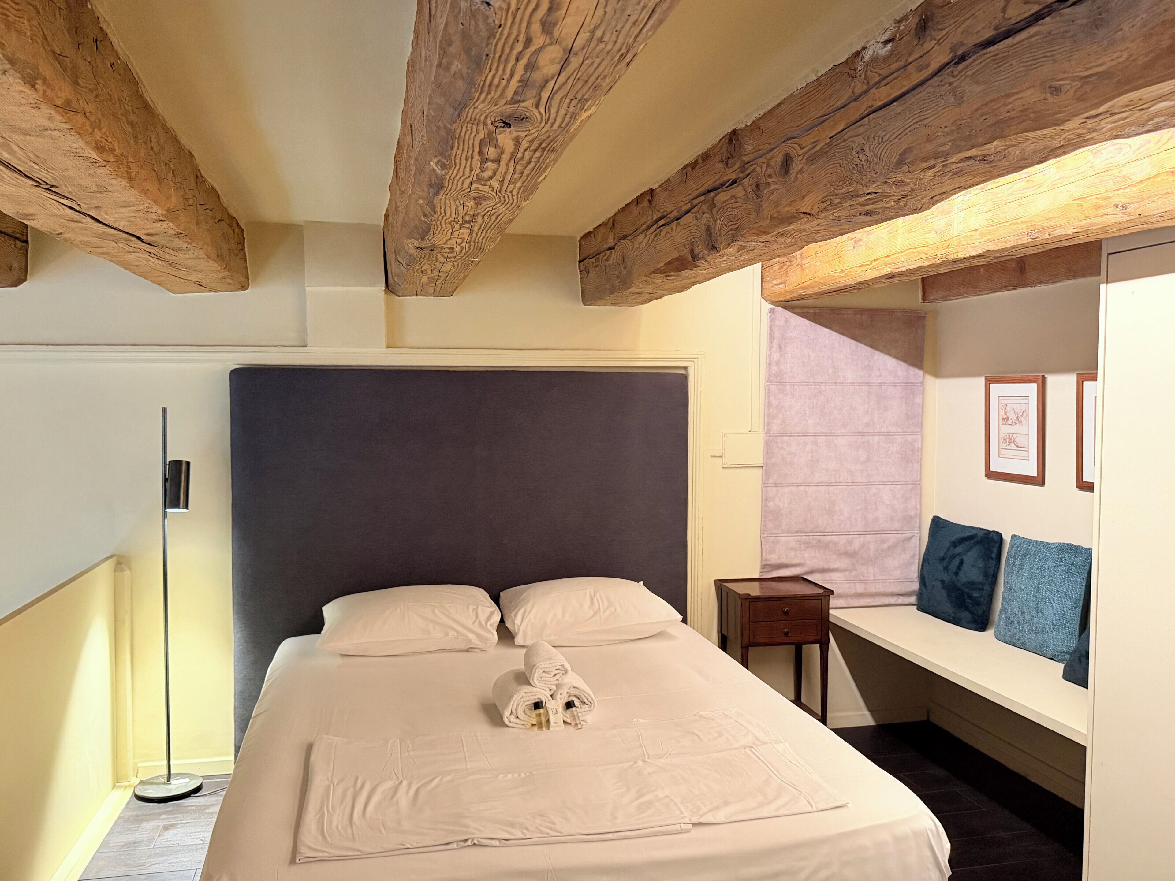 Habitación de hotel con una cama grande, vigas de madera, una mesita de noche, una lámpara y un cuadro en la pared.