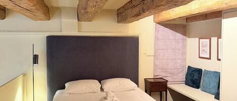 Habitación de hotel con una cama grande, vigas de madera, una mesita de noche, una lámpara y un cuadro en la pared.