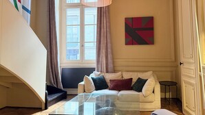 Een woonkamer met een glazen salontafel, een witte bank met kussens, een groot raam met gordijnen, een hanglamp en een muur met abstracte kunst.