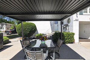 A covered patio with a glass table and chairs, a potted plant with flowers, and a white building in the background. - Midtown Reno Apartment | Near UNR & Downtown (Reno)