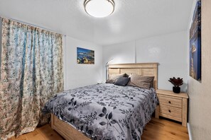A bedroom with a bed, a nightstand, a lamp, a painting, and a curtain.