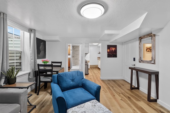 A modern living room with a blue sofa, dining table, and chairs, a staircase leading to the kitchen, and a hallway with a framed picture on the wall.