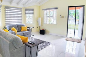 A living room with a grey sofa, yellow cushions, a coffee table, and a black ottoman. There are two windows with blinds, a door with a glass panel, and a floor-to-ceiling view of greenery. - Elegant 2BR Retreat | Trelawny, Jamaica (Trelawny Parish)