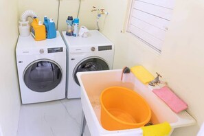 A laundry area with two washing machines, a bucket, and cleaning supplies. - Elegant 2BR Retreat | Trelawny, Jamaica (Trelawny Parish)