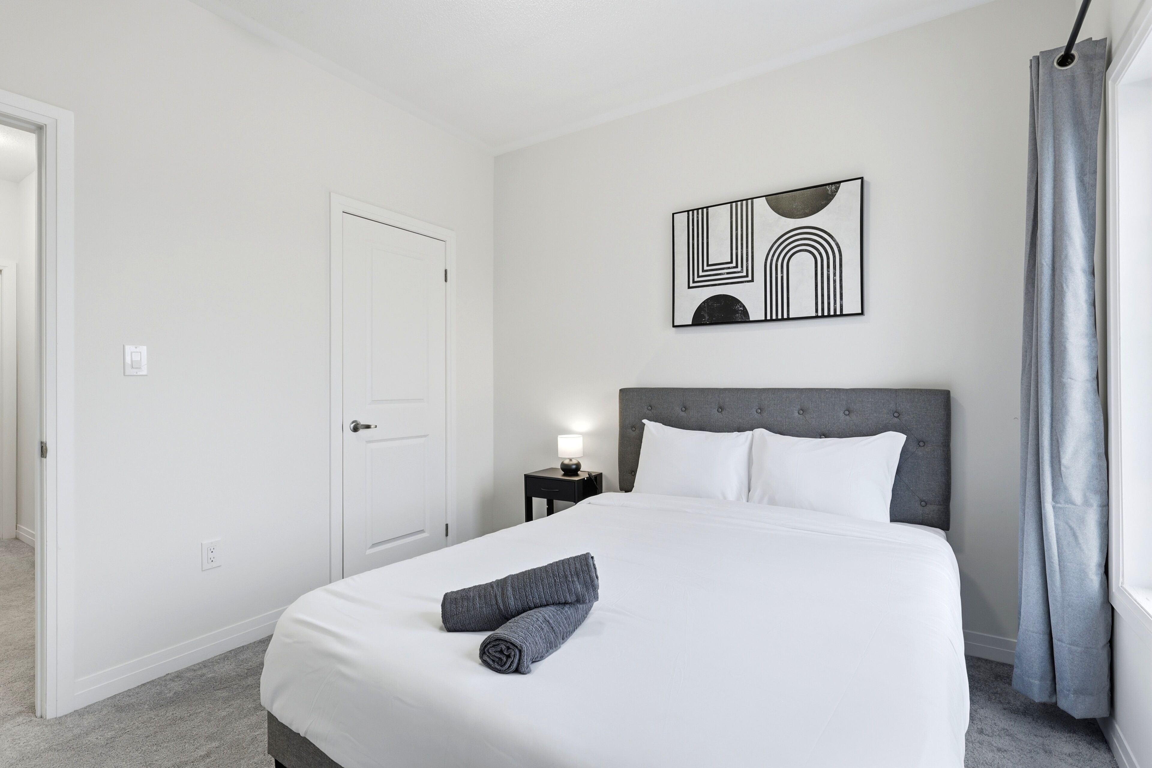 Een hotelkamer met een opgemaakt bed, een hoofdeinde, een kunstwerk aan de muur, een nachtkastje met een lamp en een raam met gordijnen.