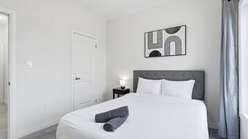 Een hotelkamer met een opgemaakt bed, een hoofdeinde, een kunstwerk aan de muur, een nachtkastje met een lamp en een raam met gordijnen.