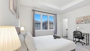 A bedroom with a bed, a desk, a chair, a window with curtains, and a lamp.
