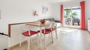Uma área de refeições com mesa de madeira e cadeiras brancas, um sofá e vista para o exterior através de uma porta de vidro deslizante.