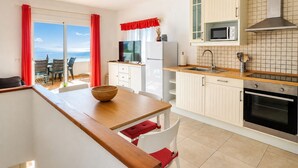 Uma cozinha com bancada de madeira, armários brancos, micro-ondas, fogão e vista para o mar através de grandes janelas.